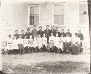 Group of students and teacher standing and sitting outside a building.