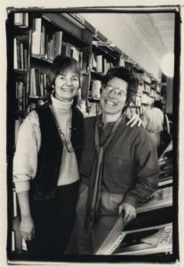 Two women facing the camera, smiling. One woman has her arm around the other woman. Bookshelves are in the background.