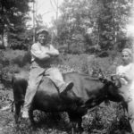 A man sitting on top of a bull in a field with another younger man holding the bull's horn.