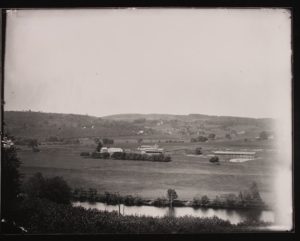 Black and white photograph of a farm with fields, buildings in the background and a river in the foreground
