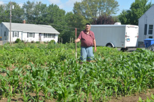 An older man standing in the middle of a large garden holding a long tool. There is a house and an equipment trailer in the background