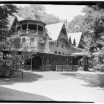 Black and white photo of a large brick and wood house. The house is asymmetrical and has many gables. There are large trees surrounding the house.