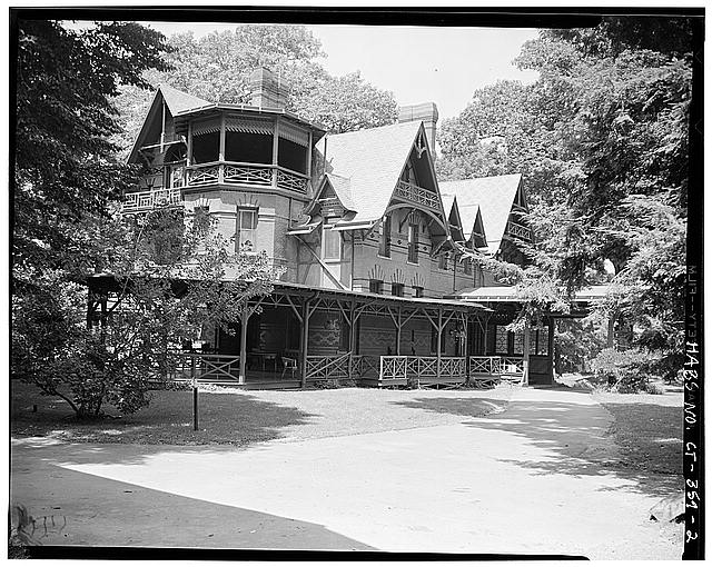 Black and white photo of a large brick and wood house. The house is asymmetrical and has many gables. There are large trees surrounding the house.