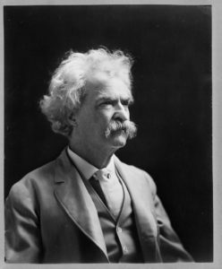 Black and white side bust portrait of a man. The man is dressed in a three piece suit and has curly hair and a bushy mustache.