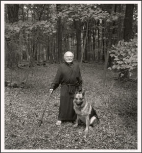 A black and white photo of an older man standing in the woods with a cane in his right hand and a german shepherd dog sitting at his feet. The man is wearing a bathrobe.