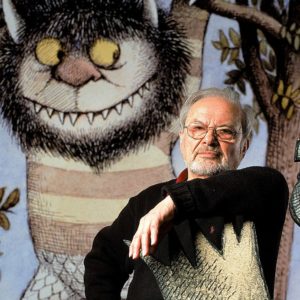 Picture of a man sitting in front of a large illustration of a monster. The man is wearing a dark sweater and has his right arm propped up.