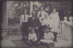 Two seated older adults--one man and one woman with five school aged children gathered around them. All facing the camera. Three children stand with their hands on the adults shoulders and two children sit on the ground in front.