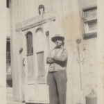 A man standing in front of a building. His arms are crossed, he's wearing a hat and smoking a pipe.