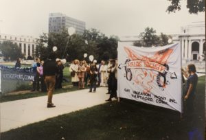 A gathering of a group of people. One person is facing the rest holding a megaphone. Two people are holding a large banner that reads "Kalos Society"