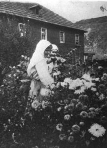 Black and white photo of a woman holding a child behind a bush of flowers. There is a large house behind them.