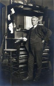 Photograph of a man standing in a three piece suit. He is leaning on a desk or cabinet and has one hand on his hip