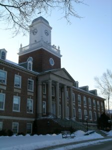 Front of a large brick building with a clock tower and columns in front of the door. There is snow on the ground.