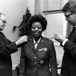 Woman in military outfit standing between two men who are pinning something to her shoulders.