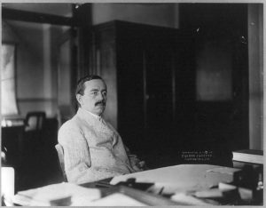 Black and white photograph of a man sitting behind a desk. He is wearing a suit jacket and has a mustache and is looking to the side of the camera