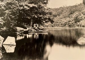 Black and white photograph of a lake surrounded by trees on the banks. There are people on the shore with a small boat