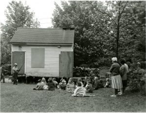 A small building jacked up. There is a group of children sitting on the ground and several adults behind the children.