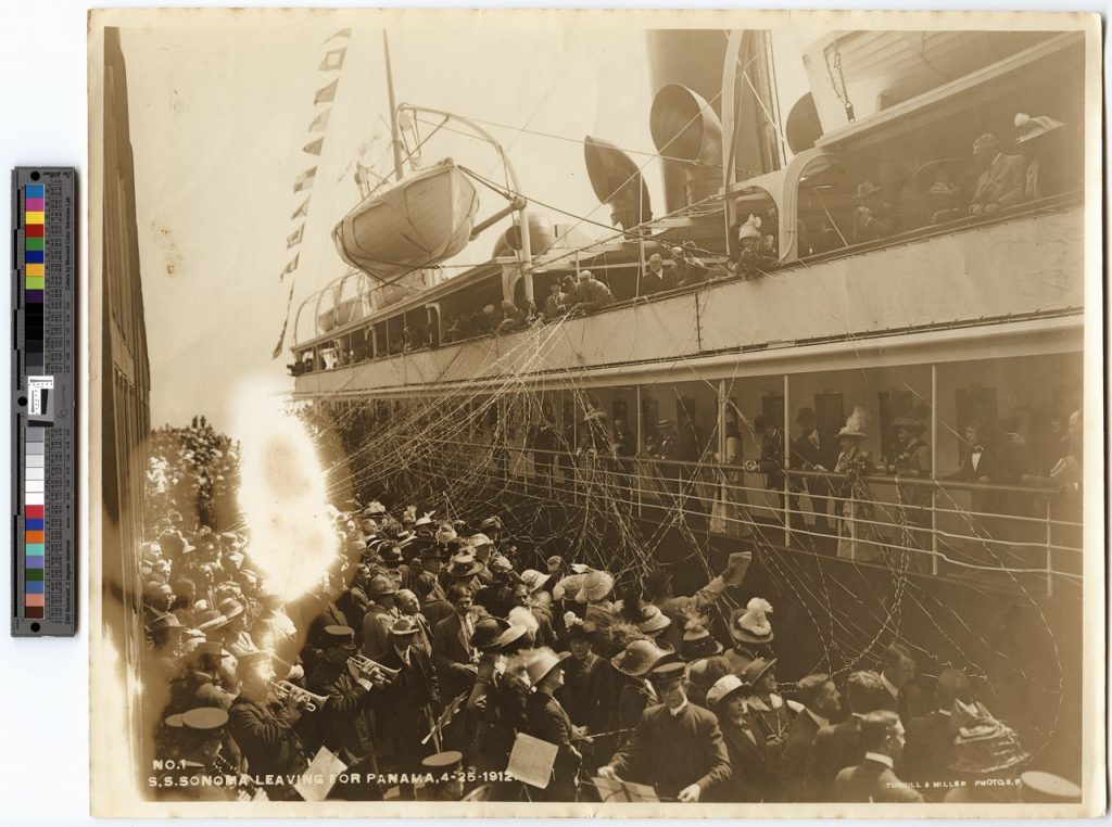 Black and white photograph of a large ship next to a dock full of hundreds of people. There are people standing on the ship and streamers coming off the boat