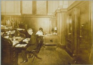 Man and woman sitting at two desks in an office. Both are looking down at something on their desk