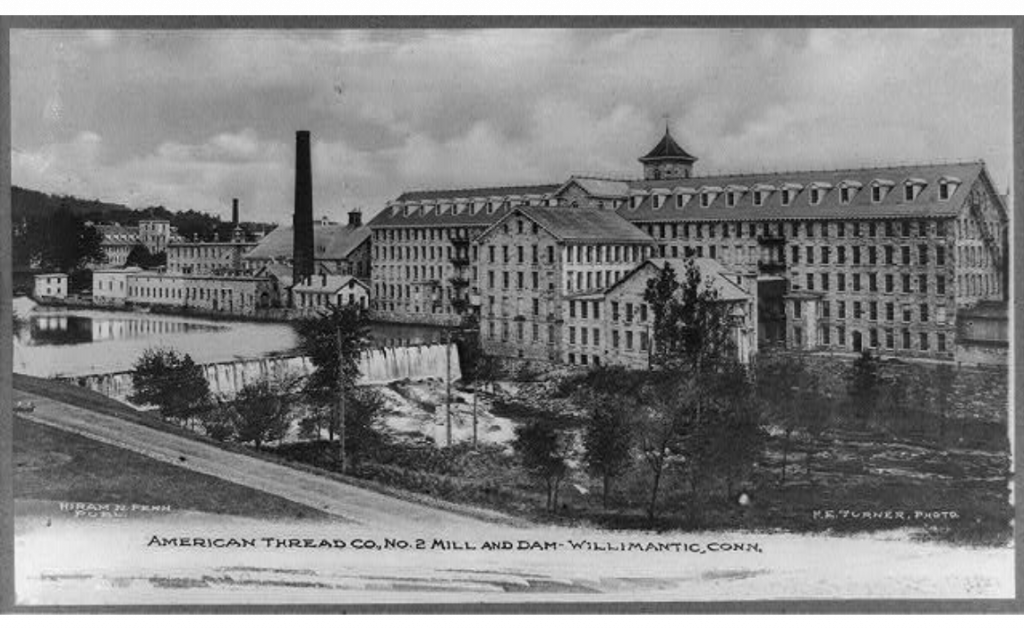 Black and white photograph of a long large building. There is a river and dam in front