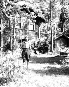 Black and white image of a man standing in front of a house and smaller building