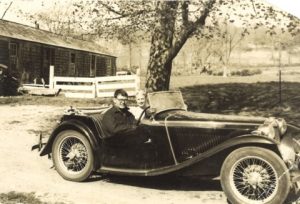 A man and a woman sitting in a black MGTC car in front of a house and a tree