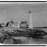 Rocky shore in front of a white lighthouse and several white buildings.