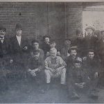Black and white photograph of a group of men sitting or standing in front of a brick building