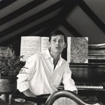 Man sitting at a piano, turned away from the piano, facing the photographer. He is wearing a white shirt. There is a potted plant to his left and lots of music books on the piano