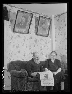 A man and woman sitting on a couch, reading a newspaper. There are two framed portraits hanging on the wall above them.