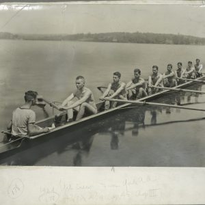 A photograph of a rowing shell with 8 rowers sitting at attention and one coxswain on the water