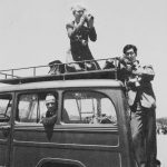 A car with one person driving and a man with a camera standing on the back bumper and a woman kneeling on the roof with a camera.