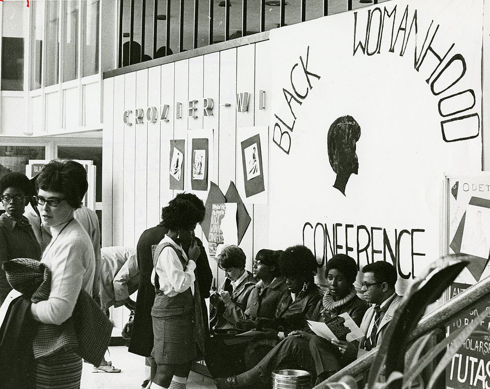 Group of women sitting and standing against a wall with a large banner reading "Black Womanhood Conference."