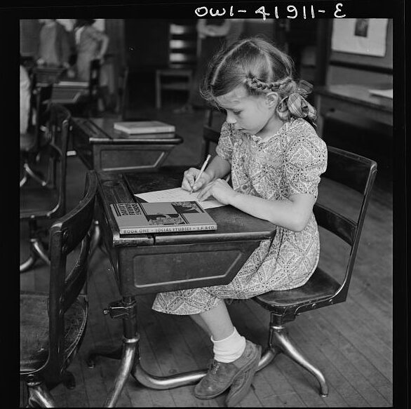 Young girl sitting at a school desk with a book, writing on a notepad or notebook.