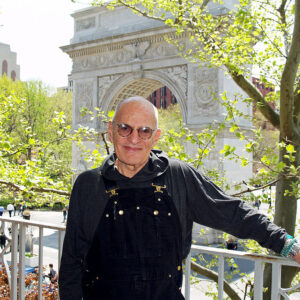 Man in a black sweatshirt and black overalls standing on a balcony.