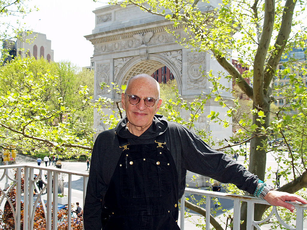 Man in a black sweatshirt and black overalls standing on a balcony.