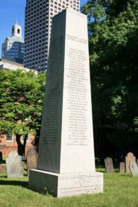 Photograph of an obelisk monument with names inscribed and other gravestones surrounding the monument.