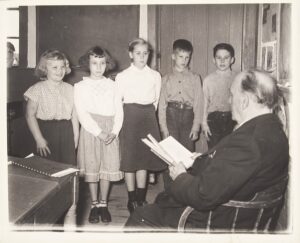 Five children standing with a seated man with a book.