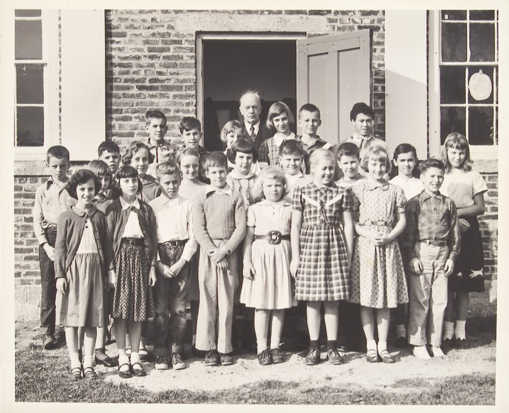 Class photograph of a group of students and male teacher standing in front of a brick building.