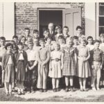 Class photograph of a group of students and male teacher standing in front of a brick building.