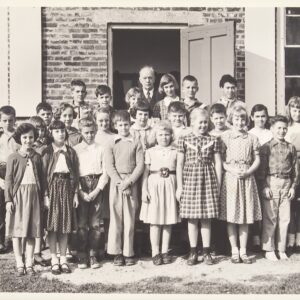 Class photograph of a group of students and male teacher standing in front of a brick building.
