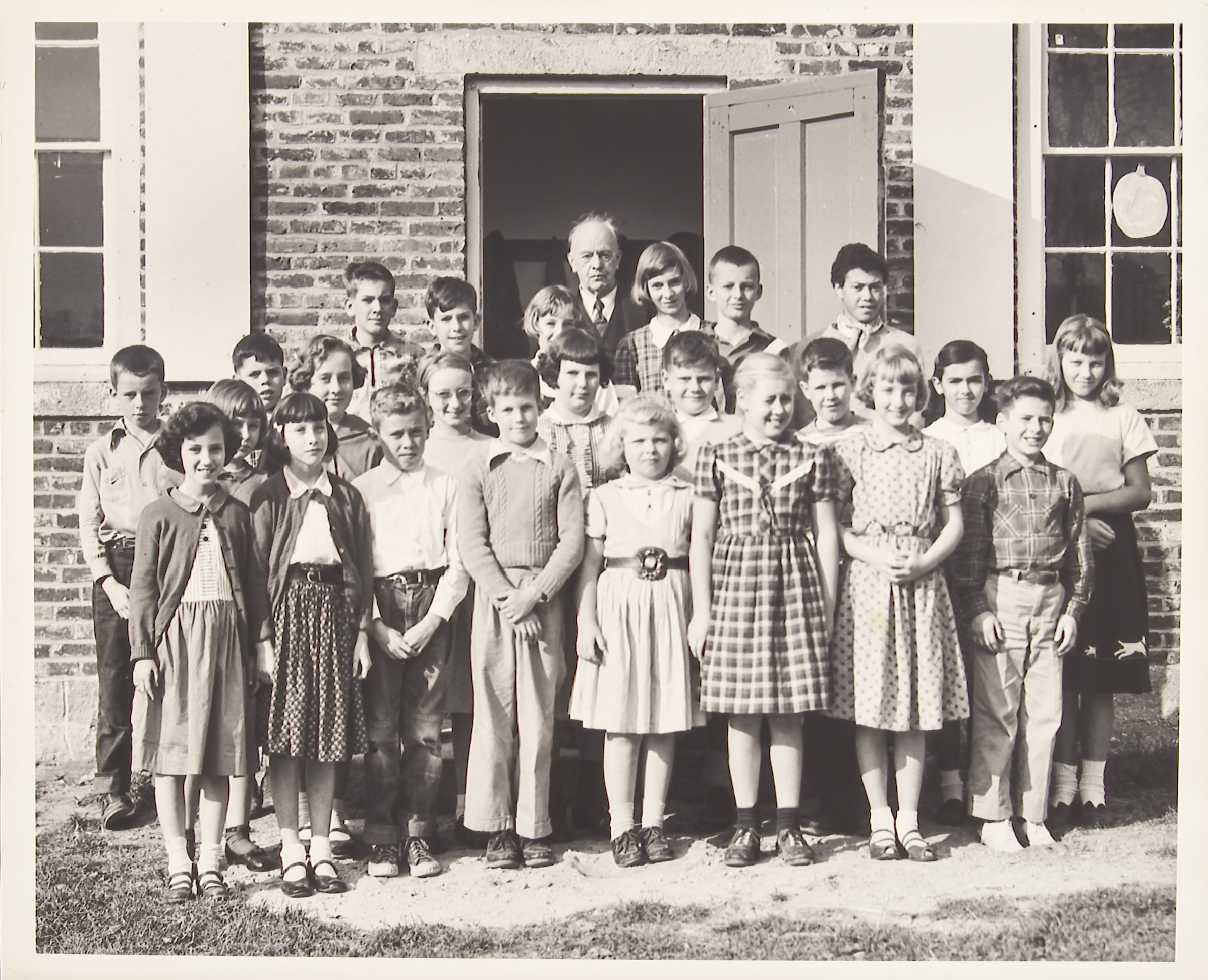 Class photograph of a group of students and male teacher standing in front of a brick building.