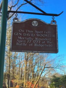 Photograph of a sign hanging along a street with the writing "On this spot fell Gen. David Wooster. Mortally wounded April 27, 1777 at the Battle of Ridgefield."