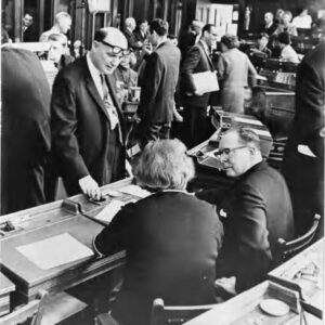 Two people seated at a table talking to one person standing. There are more people in the background.