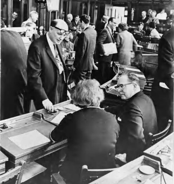 Two people seated at a table talking to one person standing. There are more people in the background.